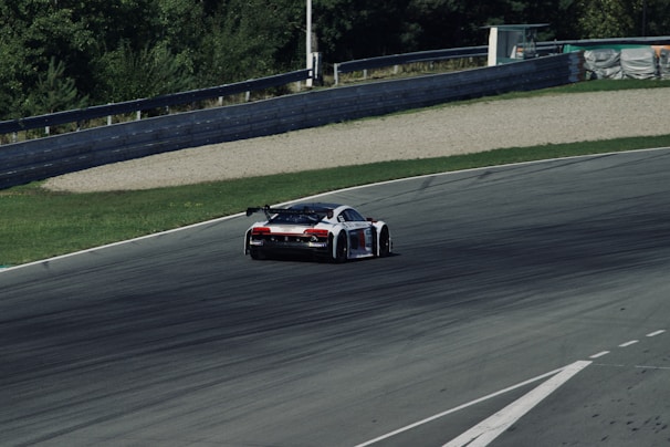 A sleek race car speeds along a curved asphalt track surrounded by greenery. The car features a sporty design with aerodynamic features and sponsorship decals. It is positioned towards the upper center of the image, leaning into the curve.