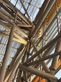 A complex arrangement of metal beams and glass panels forming a geometric structure. The beams crisscross at various angles, creating an intricate pattern. Natural light filters through the glass, casting shadows and highlighting the industrial design.