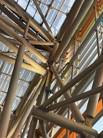 A complex arrangement of metal beams and glass panels forming a geometric structure. The beams crisscross at various angles, creating an intricate pattern. Natural light filters through the glass, casting shadows and highlighting the industrial design.