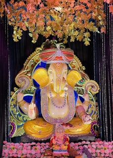 A large, ornate statue of Lord Ganesha, adorned with detailed jewelry and vibrant colors, surrounded by decorations including golden leaves and pink flowers. The statue is set against a dark background, enhancing its vivid colors and intricate design.