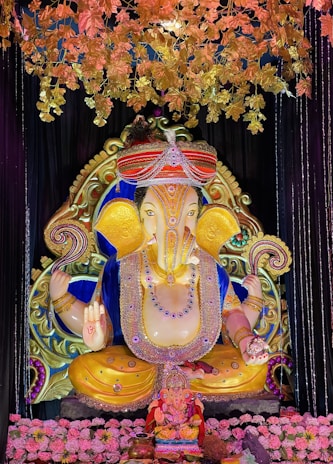A large, ornate statue of Lord Ganesha, adorned with detailed jewelry and vibrant colors, surrounded by decorations including golden leaves and pink flowers. The statue is set against a dark background, enhancing its vivid colors and intricate design.