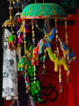 Colorful handmade fabric elephants are hanging from a green, embroidered umbrella-like structure. The elephants are adorned with beads, and small bells are attached at the bottom. In the background, there's a vibrant mix of textiles, including a red cloth with a black Om symbol and other patterned fabrics.