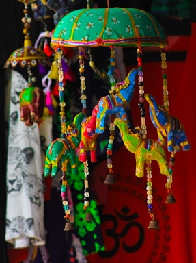 Colorful handmade fabric elephants are hanging from a green, embroidered umbrella-like structure. The elephants are adorned with beads, and small bells are attached at the bottom. In the background, there's a vibrant mix of textiles, including a red cloth with a black Om symbol and other patterned fabrics.