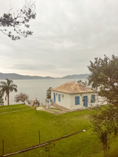 A small white house with a tiled roof and blue shutters is surrounded by green grass and trees. In the background, a calm body of water reflects a cloudy sky, with distant mountains visible on the horizon. A palm tree and other vegetation are nearby, adding to the serene setting.