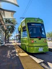 A bright green metro train is stationed at a platform, with a blue sky overhead. The platform is paved with bricks and features a textured yellow line for safety. A modern canopy provides shade, and there are trees and buildings in the background.