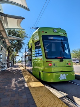 A bright green metro train is stationed at a platform, with a blue sky overhead. The platform is paved with bricks and features a textured yellow line for safety. A modern canopy provides shade, and there are trees and buildings in the background.