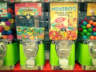 Three candy vending machines are filled with colorful candies. The middle machine features Mike and Ike candies, the right machine has Miniboo chewy sweets, and the left machine contains multicolored bouncing balls resembling soccer balls.