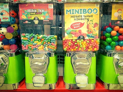 Three candy vending machines are filled with colorful candies. The middle machine features Mike and Ike candies, the right machine has Miniboo chewy sweets, and the left machine contains multicolored bouncing balls resembling soccer balls.