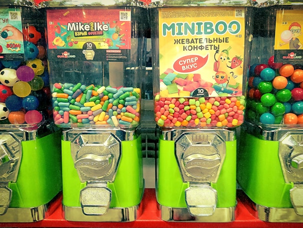 Three candy vending machines are filled with colorful candies. The middle machine features Mike and Ike candies, the right machine has Miniboo chewy sweets, and the left machine contains multicolored bouncing balls resembling soccer balls.