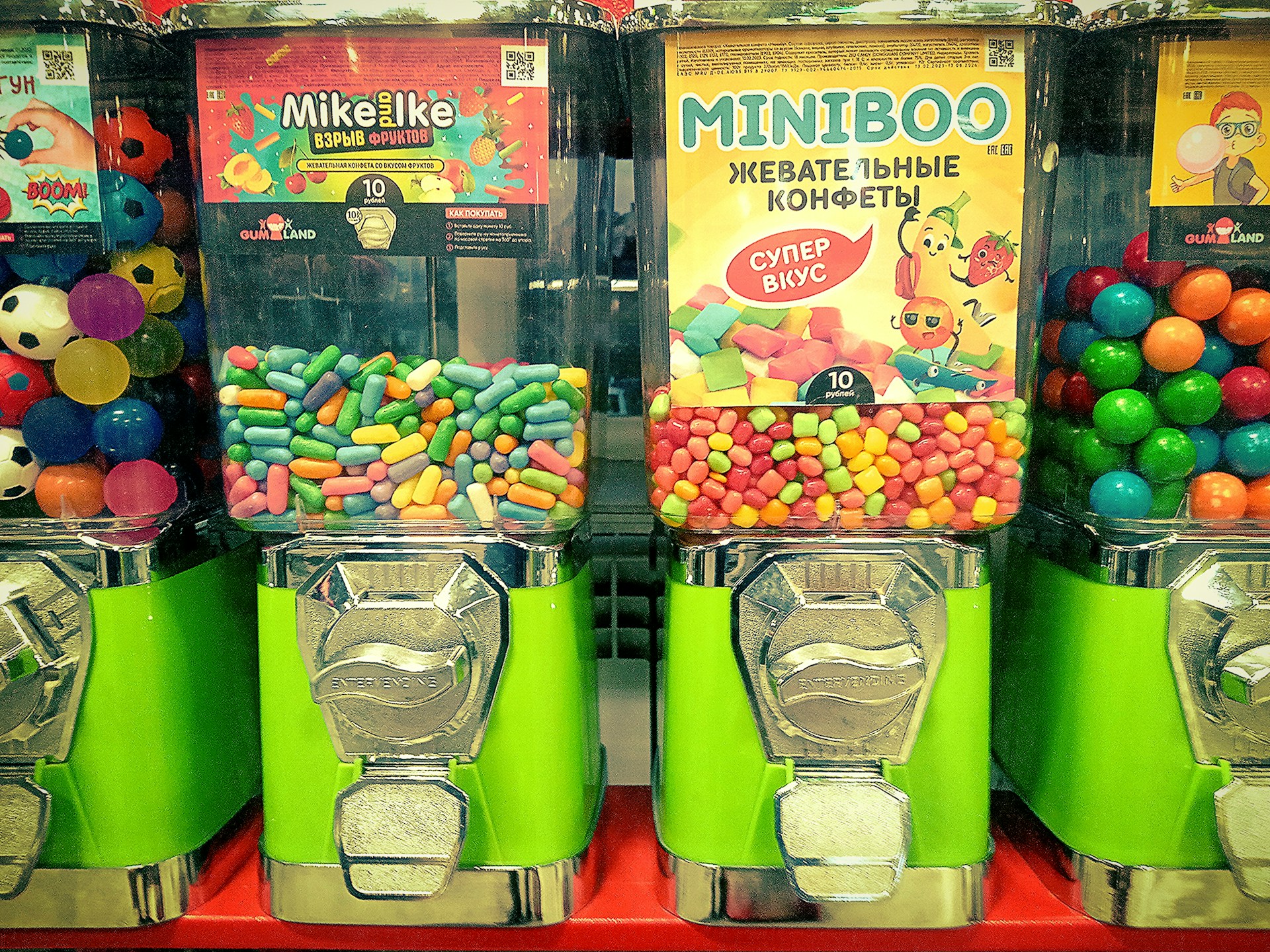 Three candy vending machines are filled with colorful candies. The middle machine features Mike and Ike candies, the right machine has Miniboo chewy sweets, and the left machine contains multicolored bouncing balls resembling soccer balls.