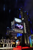 A large, illuminated building at night with the words 'Studio City' prominently displayed. The structure is adorned with bright lights and architectural designs that highlight its grandeur. A large screen above displays an advertisement or video, adding to the lively atmosphere. Surrounding the entrance are palm trees, colorful spotlights, and art deco elements.