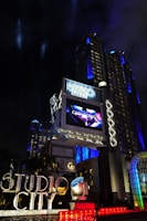 A large, illuminated building at night with the words 'Studio City' prominently displayed. The structure is adorned with bright lights and architectural designs that highlight its grandeur. A large screen above displays an advertisement or video, adding to the lively atmosphere. Surrounding the entrance are palm trees, colorful spotlights, and art deco elements.