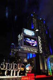 A large, illuminated building at night with the words 'Studio City' prominently displayed. The structure is adorned with bright lights and architectural designs that highlight its grandeur. A large screen above displays an advertisement or video, adding to the lively atmosphere. Surrounding the entrance are palm trees, colorful spotlights, and art deco elements.