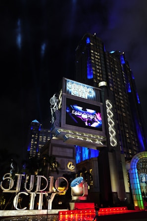 A large, illuminated building at night with the words 'Studio City' prominently displayed. The structure is adorned with bright lights and architectural designs that highlight its grandeur. A large screen above displays an advertisement or video, adding to the lively atmosphere. Surrounding the entrance are palm trees, colorful spotlights, and art deco elements.