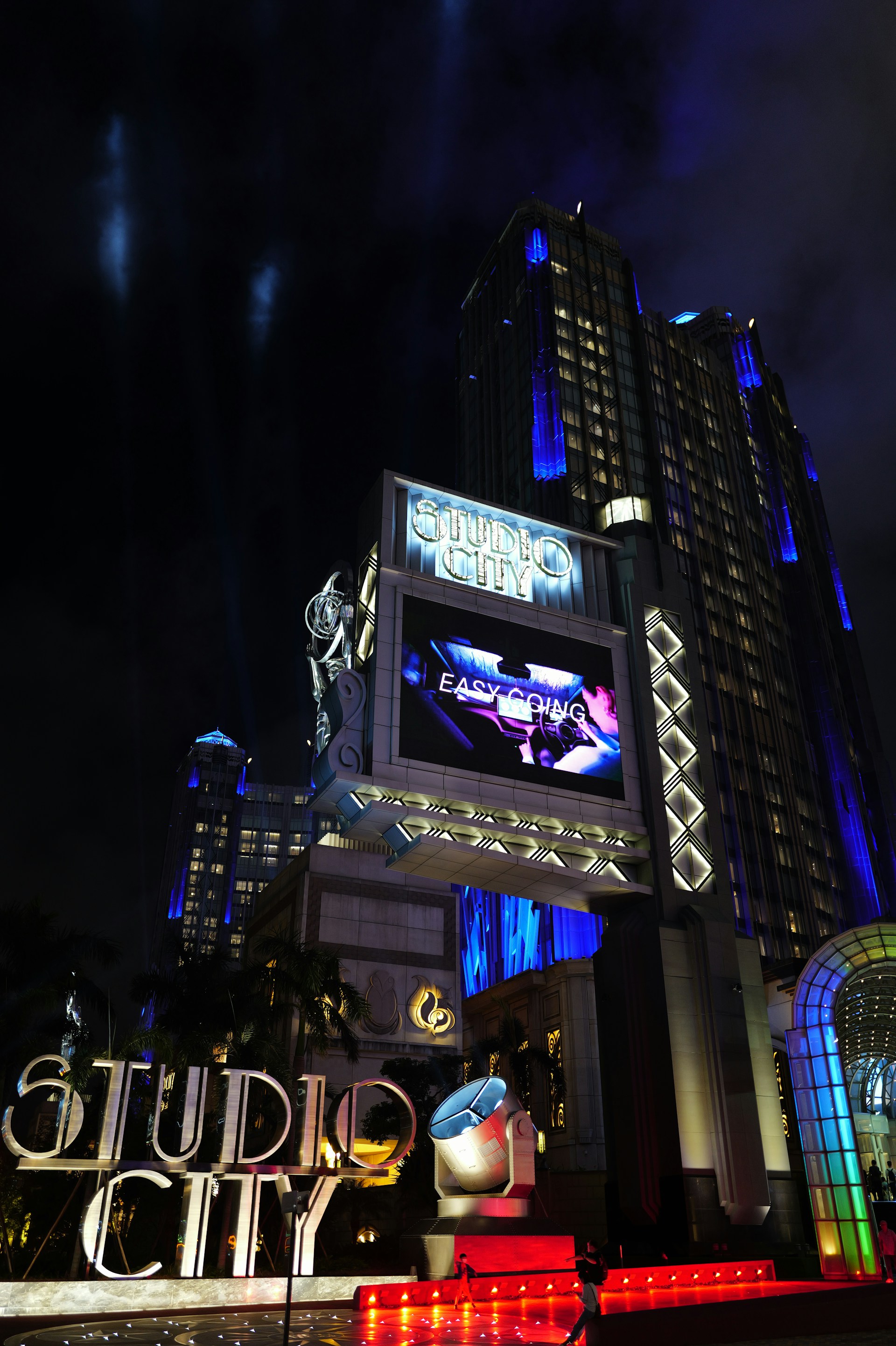 A large, illuminated building at night with the words 'Studio City' prominently displayed. The structure is adorned with bright lights and architectural designs that highlight its grandeur. A large screen above displays an advertisement or video, adding to the lively atmosphere. Surrounding the entrance are palm trees, colorful spotlights, and art deco elements.