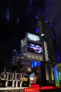 A large, illuminated building at night with the words 'Studio City' prominently displayed. The structure is adorned with bright lights and architectural designs that highlight its grandeur. A large screen above displays an advertisement or video, adding to the lively atmosphere. Surrounding the entrance are palm trees, colorful spotlights, and art deco elements.