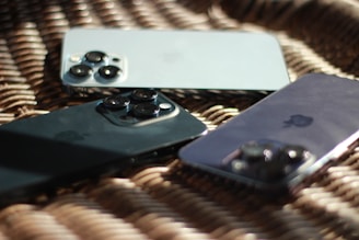 Three smartphones lie on a woven rattan surface, each displaying a reflective back with distinct triple-camera setups placed diagonally. The lighting creates a soft glow on the devices, emphasizing their sleek metallic finishes.