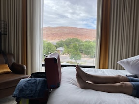 A person is lying on a bed inside a hotel room with an open suitcase nearby. Through the window, there is a view of a red rock hill and some greenery. The window also shows a street with vehicles.
