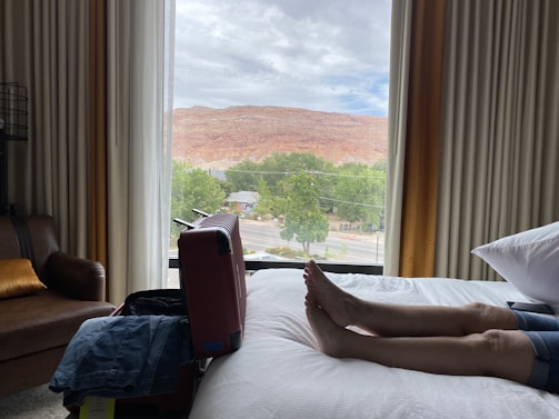 A person is lying on a bed inside a hotel room with an open suitcase nearby. Through the window, there is a view of a red rock hill and some greenery. The window also shows a street with vehicles.