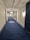A long corridor with blue carpet flooring, white walls, and dark blue doors. The ceiling is exposed with pipes and light fixtures. A patterned design is visible on the carpet in the distance.