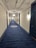 A long corridor with blue carpet flooring, white walls, and dark blue doors. The ceiling is exposed with pipes and light fixtures. A patterned design is visible on the carpet in the distance.