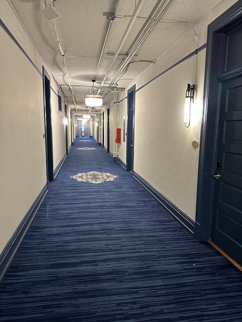 A long corridor with blue carpet flooring, white walls, and dark blue doors. The ceiling is exposed with pipes and light fixtures. A patterned design is visible on the carpet in the distance.