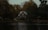 A quaint house with a red facade and white trim is nestled among tall, dense trees with autumn foliage. It is situated by a calm, reflective body of water, creating a serene and picturesque scene.