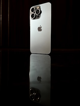 A modern smartphone with a sleek design and multiple camera lenses on the back is positioned upright on a reflective surface, creating a mirror image. The logo of the brand is visible in the center of the phone's back. The background is dark, which highlights the phone's metallic color.