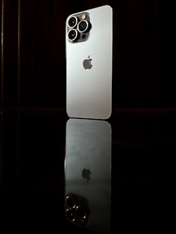 A modern smartphone with a sleek design and multiple camera lenses on the back is positioned upright on a reflective surface, creating a mirror image. The logo of the brand is visible in the center of the phone's back. The background is dark, which highlights the phone's metallic color.