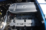 A detailed view of a car engine, prominently featuring a Ford Racing 427 Cobra engine with visible components such as the air filter, valves, and spark plug wires. The engine is clean and well-maintained, with metallic and mechanical parts showing intricate detailing.