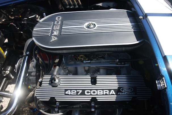 A detailed view of a car engine, prominently featuring a Ford Racing 427 Cobra engine with visible components such as the air filter, valves, and spark plug wires. The engine is clean and well-maintained, with metallic and mechanical parts showing intricate detailing.