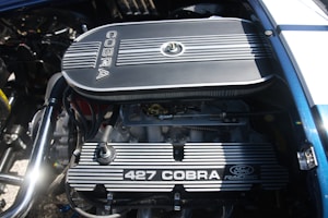A detailed view of a car engine, prominently featuring a Ford Racing 427 Cobra engine with visible components such as the air filter, valves, and spark plug wires. The engine is clean and well-maintained, with metallic and mechanical parts showing intricate detailing.