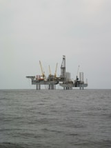 An offshore oil rig is situated in the middle of the sea, with multiple cranes and a complex structure made up of platforms and supports. The surrounding water appears calm, and the sky is overcast, giving a sense of isolation and vastness.