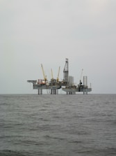 An offshore oil rig is situated in the middle of the sea, with multiple cranes and a complex structure made up of platforms and supports. The surrounding water appears calm, and the sky is overcast, giving a sense of isolation and vastness.