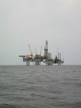 An offshore oil rig is situated in the middle of the sea, with multiple cranes and a complex structure made up of platforms and supports. The surrounding water appears calm, and the sky is overcast, giving a sense of isolation and vastness.