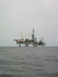 An offshore oil rig is situated in the middle of the sea, with multiple cranes and a complex structure made up of platforms and supports. The surrounding water appears calm, and the sky is overcast, giving a sense of isolation and vastness.