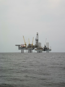 An offshore oil rig is situated in the middle of the sea, with multiple cranes and a complex structure made up of platforms and supports. The surrounding water appears calm, and the sky is overcast, giving a sense of isolation and vastness.