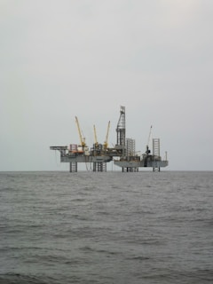 An offshore oil rig is situated in the middle of the sea, with multiple cranes and a complex structure made up of platforms and supports. The surrounding water appears calm, and the sky is overcast, giving a sense of isolation and vastness.