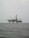 An offshore oil rig is situated in the middle of the sea, with multiple cranes and a complex structure made up of platforms and supports. The surrounding water appears calm, and the sky is overcast, giving a sense of isolation and vastness.
