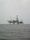 An offshore oil rig is situated in the middle of the sea, with multiple cranes and a complex structure made up of platforms and supports. The surrounding water appears calm, and the sky is overcast, giving a sense of isolation and vastness.
