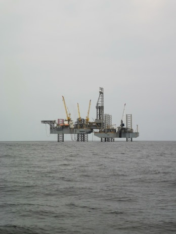 An offshore oil rig is situated in the middle of the sea, with multiple cranes and a complex structure made up of platforms and supports. The surrounding water appears calm, and the sky is overcast, giving a sense of isolation and vastness.