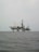An offshore oil rig is situated in the middle of the sea, with multiple cranes and a complex structure made up of platforms and supports. The surrounding water appears calm, and the sky is overcast, giving a sense of isolation and vastness.