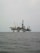 An offshore oil rig is situated in the middle of the sea, with multiple cranes and a complex structure made up of platforms and supports. The surrounding water appears calm, and the sky is overcast, giving a sense of isolation and vastness.