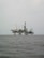 An offshore oil rig is situated in the middle of the sea, with multiple cranes and a complex structure made up of platforms and supports. The surrounding water appears calm, and the sky is overcast, giving a sense of isolation and vastness.