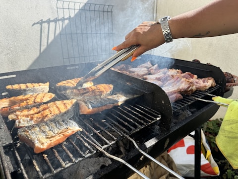 A grill loaded with fish and meat is being tended to with metal tongs. The food is cooking over charcoal, emitting smoke and grill marks are visible on the food. A person's hand is adjusting the food on the grill, wearing a watch and with long painted nails.