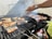 A grill loaded with fish and meat is being tended to with metal tongs. The food is cooking over charcoal, emitting smoke and grill marks are visible on the food. A person's hand is adjusting the food on the grill, wearing a watch and with long painted nails.