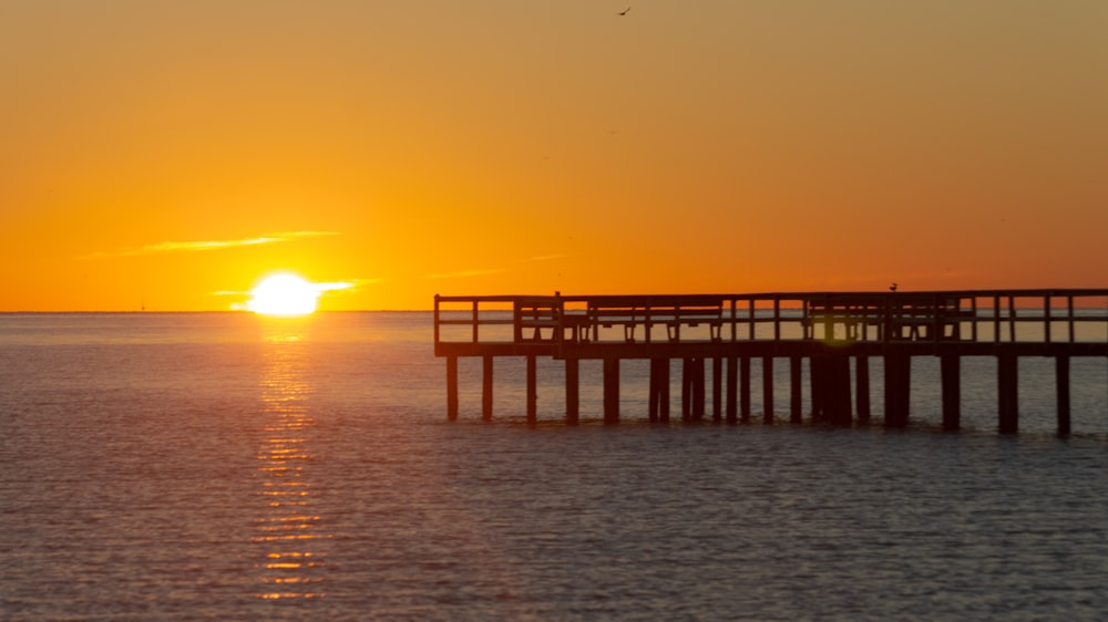 the sun is setting over the ocean with a pier in the foreground