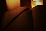 A candle lit beside an open book with elegant typography, casting gentle shadows on a cream-colored surface.