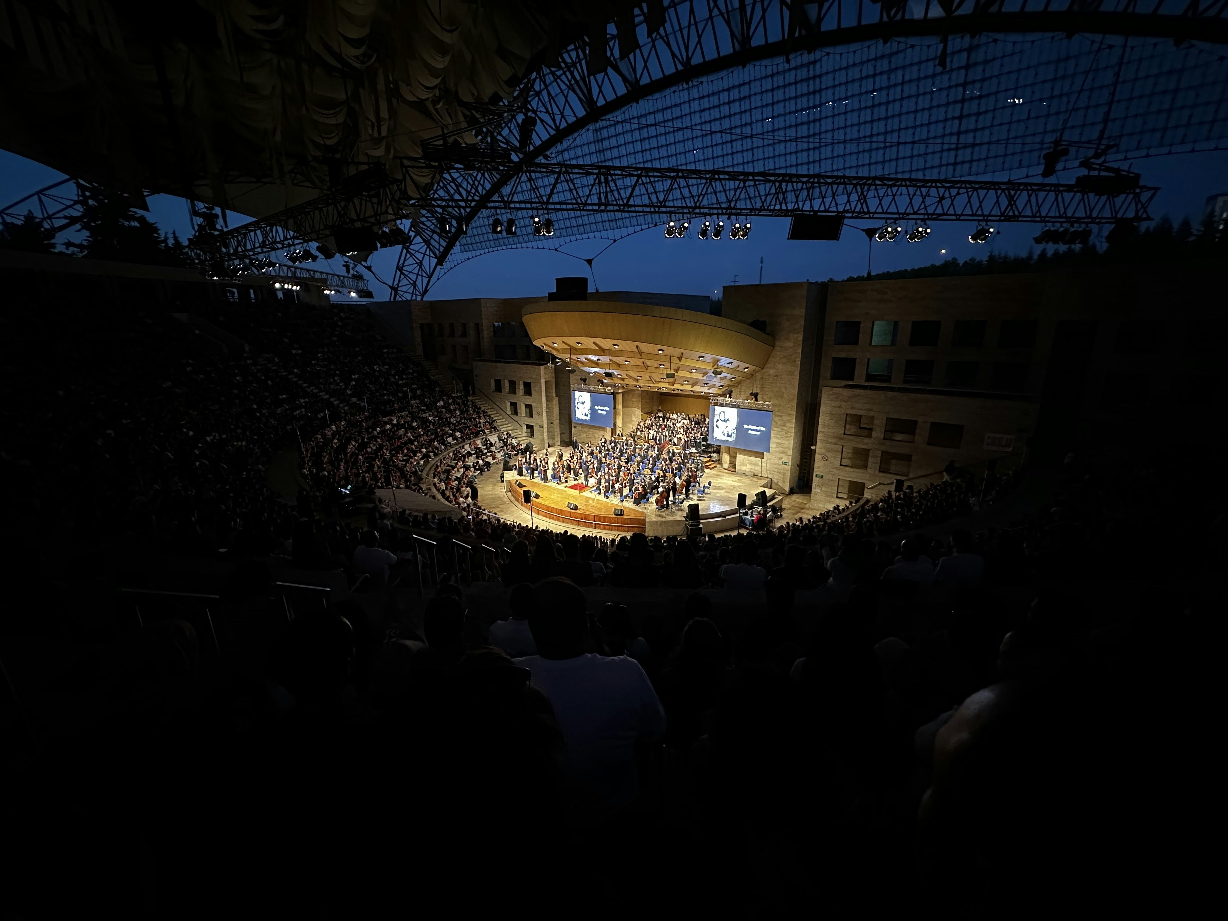 Symphony orchestra performing under stars at outdoor amphitheater, warm stage lighting, dark evening sky