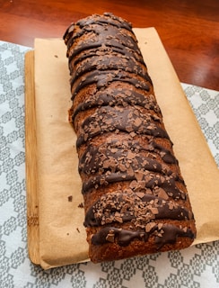 A loaf-shaped cake with a rich chocolate glaze and chocolate shavings, placed on a wooden board covered with parchment paper. The cake is resting on a patterned fabric surface.
